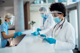Black female doctor using digital tablet while standing at reception desk at medical clinic and wearing face shield and mask due to COVID-19 pandemic.