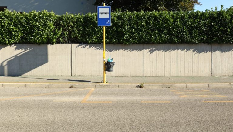 Bus stop with pole, sign and full waste bin. Concrete wall and hedge on behind, asphalt road in front.