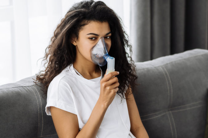 Ill African american woman with an inhaler, sick millennial girl doing inhalation at home, she use nebulizer and inhaler for the treatment sitting on the couch at home at quarantine