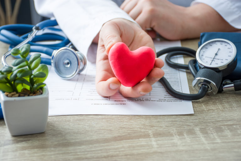 Doctor of internal medicine and cardiologist holding in his hands and shows to patient figure of red card heart during medical consultation. Explanation of causes of heart, diagnosis and treatment