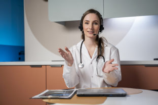 Young female doctor in a white medical uniform with a stethoscope, using a computer laptop, talks via videoconference with a patient, looking into the camera at a healthcare hospital
