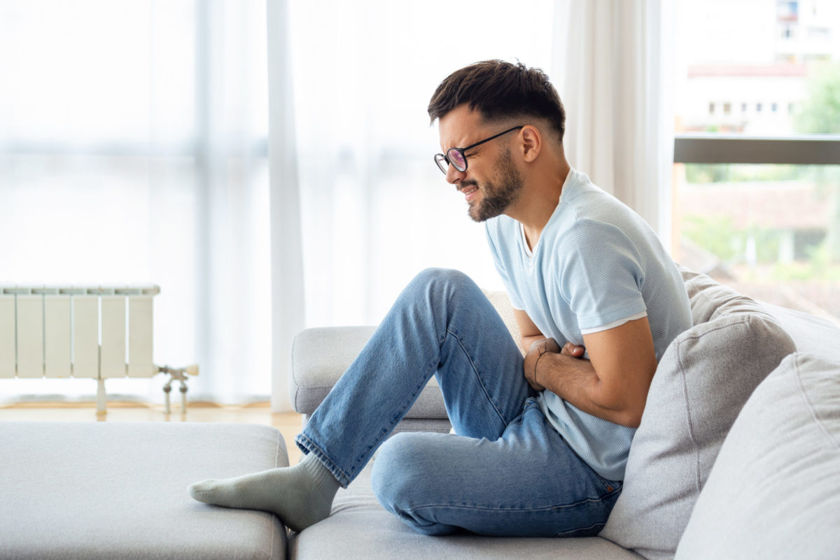 A man suffers from abdominal pain while sitting at home on the couch. Young man suffering from abdominal pain sitting on the couch at home.