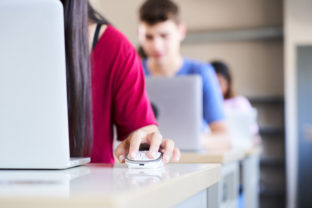 Close up of a hand clutching a mouse in a high school classroom, a group of cheerful students study with a laptop computer. Concept of technology and education