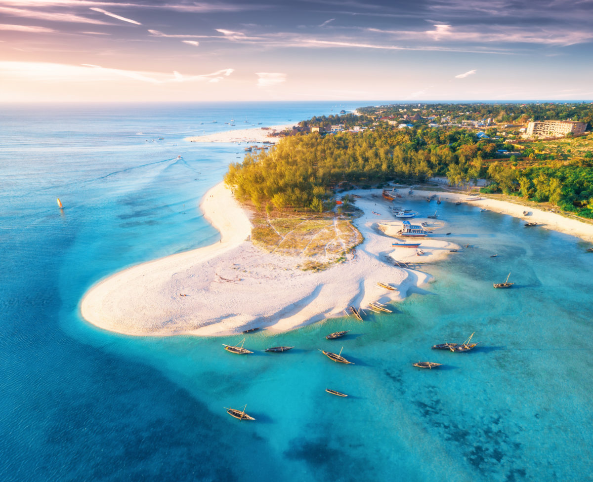 Aerial view of the fishing boats on tropical sea coast with sandy beach at sunset. Summer travel in Zanzibar, Africa. Top view of boats, yachts, green palm trees, clear blue water, colorful sky