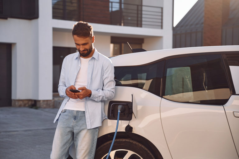 Charging the vehicle near the house. Holding smartphone. Young stylish man is with electric car at daytime