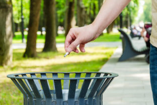 Closeup of female hand throwing cigarette butt into a metal trash can in a city park.
