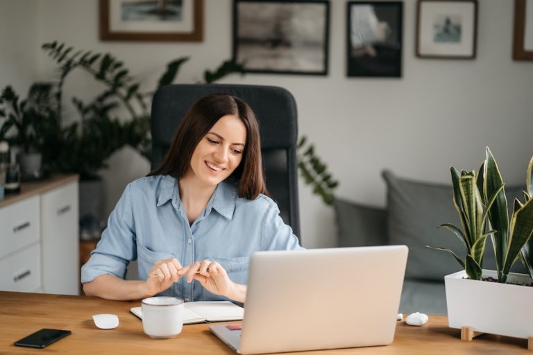 Young pretty woman talking on video call and waving hand while sitting at table in the homeoffice room
