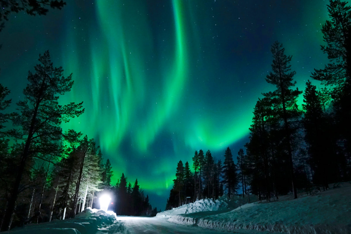 Bright lamp shining on the empty snowy road just as the northern lights appear.