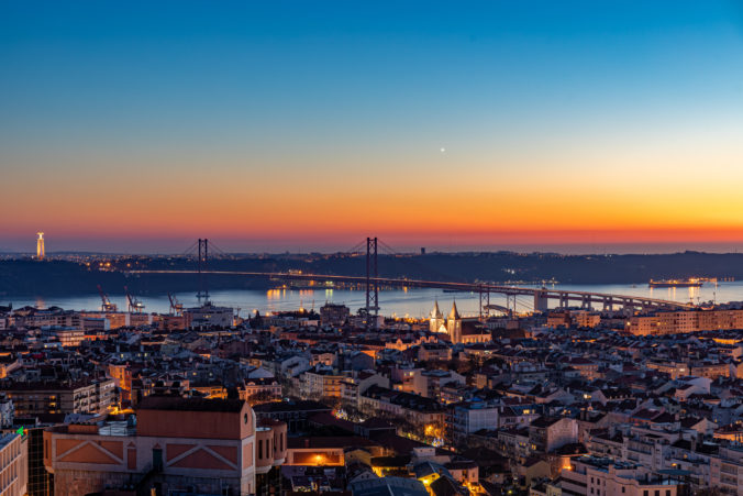 Aerial view of the cityscape of Lisbon with illuminated buildings, bridge, and sea during the sunset