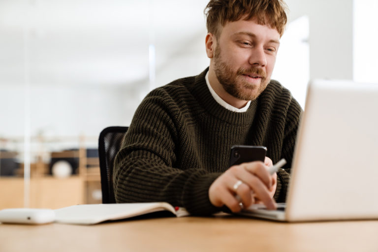 Young ginger man working with mobile phone and laptop in office
