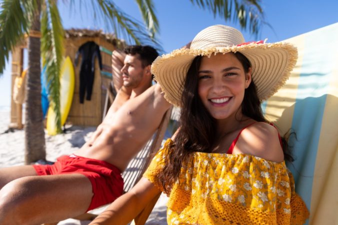 Caucasian couple enjoying time at the beach, sitting on deck chairs, a man is sunbathing and a woman is looking at the camera and smiling