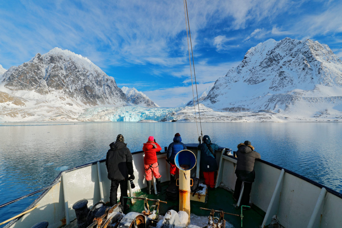 Holiday travel in Arctic, Svalbard, Norway. People on the boat. Winter mountain with snow, blue glacier ice with sea in the foreground. Blue sky with white clouds. Snowy hill in ocean. Travel in sea.
