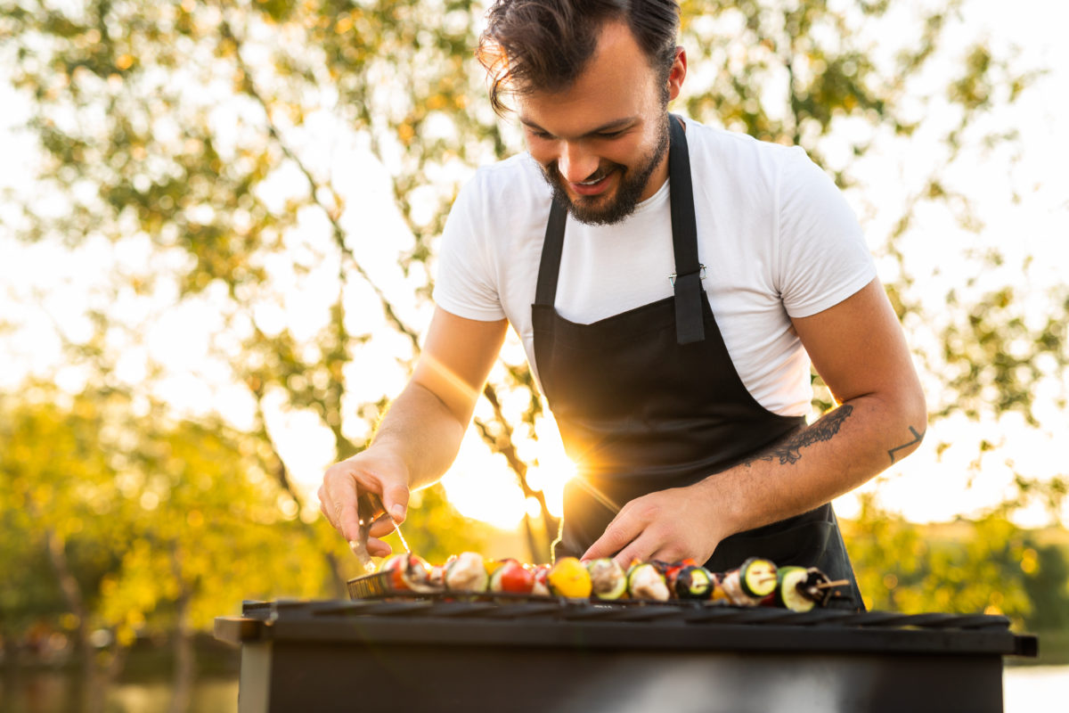 Joyful ethnic man grilling vegetable kebabs in park