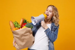 Cheerful woman in denim clothes isolated on orange background. Delivery service from shop or restaurant concept. Hold brown craft paper bag for takeaway mock up with food products scream in megaphone.