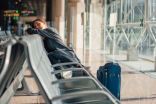 Young tired woman sleeping alone in empty airport with her hand luggage, waiting flight - transportation, low cost traveling, delayed or cancelled flight concept