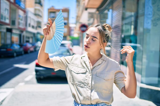 Young beautiful blonde caucasian woman smiling happy outdoors on a sunny day using handfan for hot weather