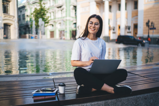 Carefree female student in classic eyeglasses thoughtful looking away and dreaming while e learning on distance using laptop computer during digital travelling, happy pondering girl with netbook