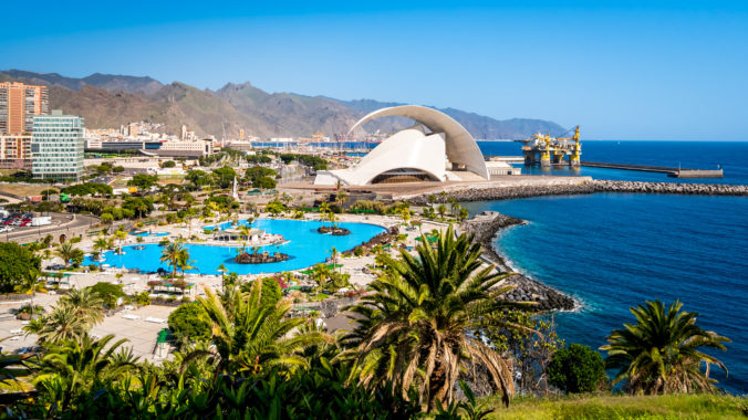 Santa Cruz de Tenerife with iconic Auditorio de Tenerife concert hall shining in the sunlight against a backdrop of Anaga mountains, an oil rig and a tropical pool with palm trees in the foreground.