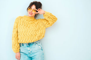 Young beautiful smiling female in trendy summer yellow hipster sweater and  jeans. Sexy carefree woman posing near light blue wall in studio. Positive model having fun indoors.Shows peace sign
