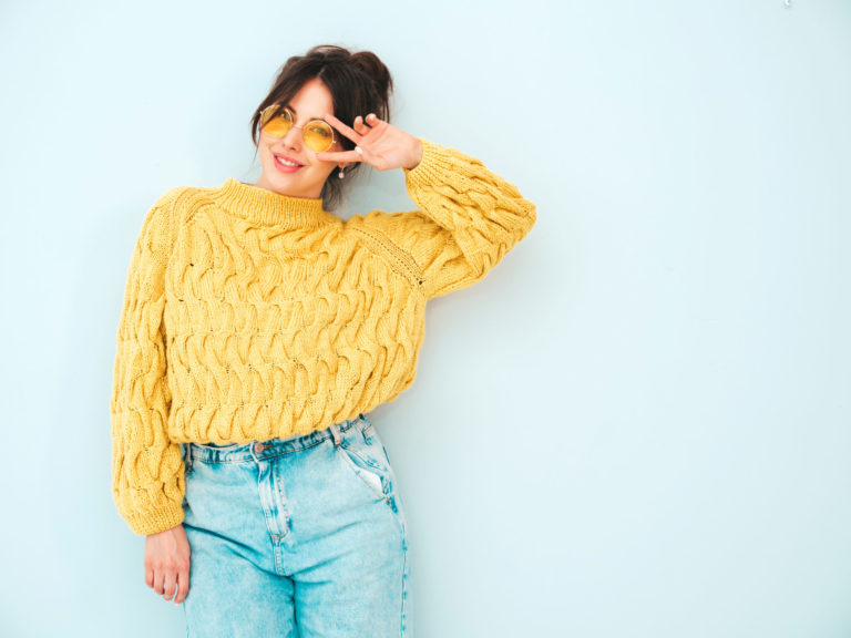 Young beautiful smiling female in trendy summer yellow hipster sweater and  jeans. Sexy carefree woman posing near light blue wall in studio. Positive model having fun indoors.Shows peace sign