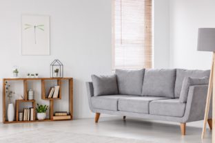 Real photo of a scandi living room interior with gray settee standing near the window, next to a wooden bookcase with plants and books