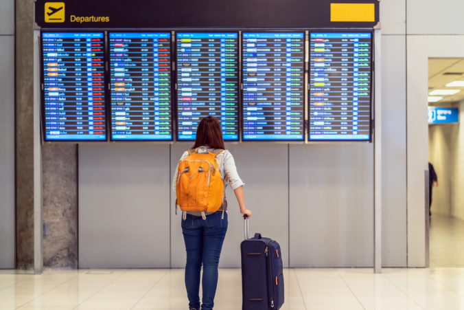 Back side of traveler with luggage standing over the flight board for check in at the flight information screen in modern an airport, travel and transportation with technology concept.