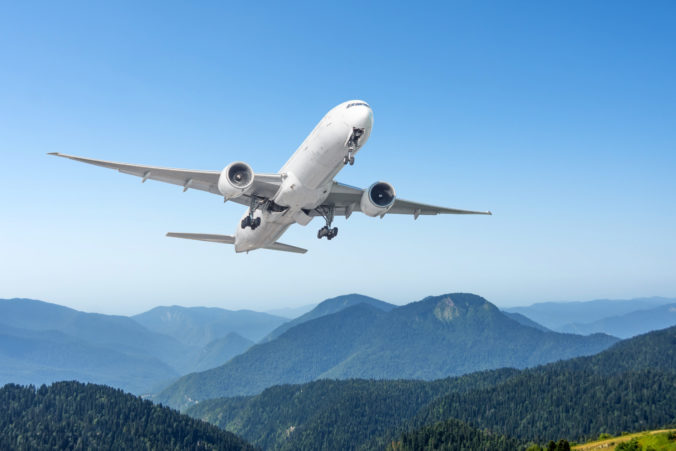 Commercial passenger airliner with extended landing gear flies over mountain rocky hilly landscape on a clear sunny day.