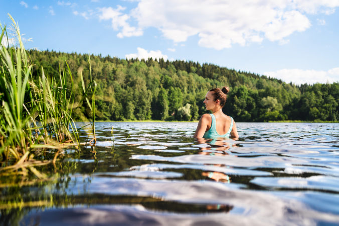 Swimming in lake in Finland. Woman in summer at beach. Finnish bathing in water in nature. Back view of person. Blue sky and beautiful green forest.