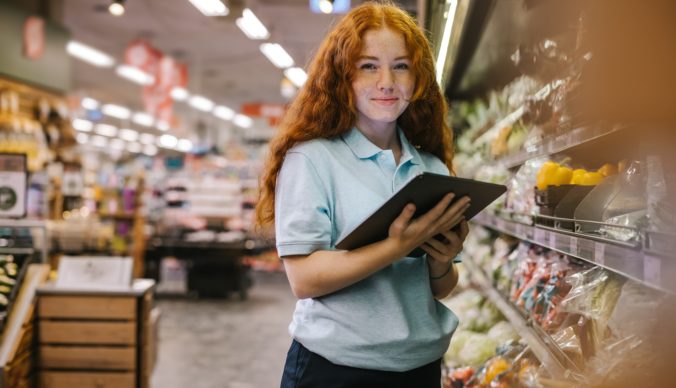 Alt text: Tínedžerka s dlhými ryšavými vlasmi drží tablet a usmieva sa, stojí v uličke potravinového obchodu plnej čerstvej zeleniny.