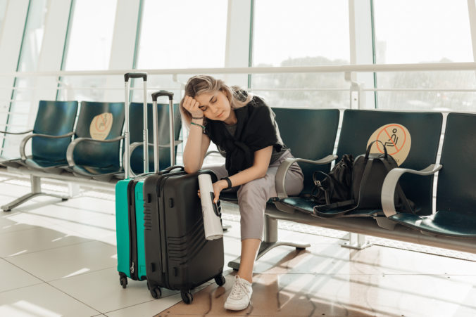 Bored blond woman with luggage, leaning elbow on bags, sit in waiting room at airport due to Covid travel restriction