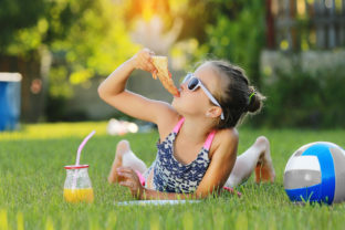 Kid girl is eating piece of pizza and playing on laptop lying on grass on summer sunny day. the child has lunch after active games. hammock and ball on the background.