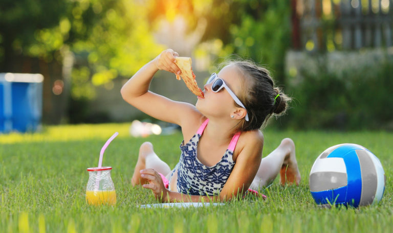 Kid girl is eating piece of pizza and playing on laptop lying on grass on summer sunny day. the child has lunch after active games. hammock and ball on the background.