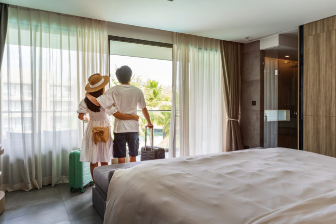 Young couple traveler opening the curtains and looking at the view from the window of a hotel room while on summer vacation, Travel lifestyle concept