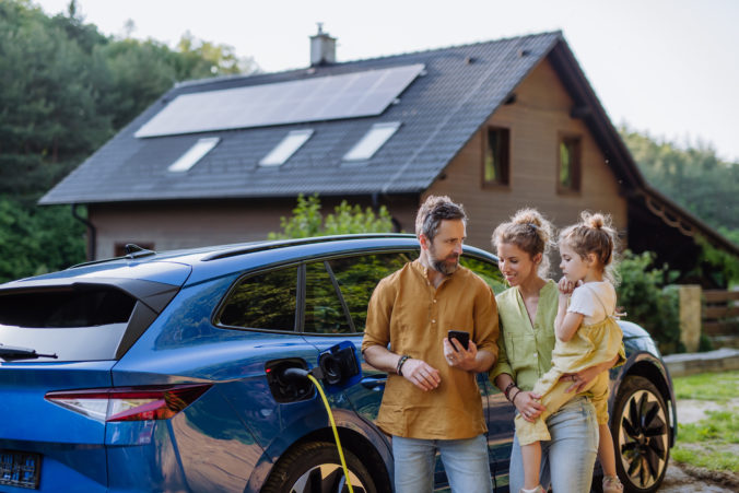 Family with little girl standing in front of their house with solar panels on the roof, having electric car.