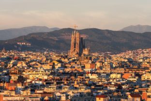 Panoramic view of Barcelona with Sagrada Familia in the center, surrounded by city buildings and mountains in the background.