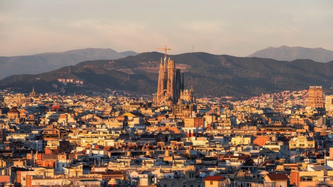 Panoramic view of Barcelona with Sagrada Familia in the center, surrounded by city buildings and mountains in the background.