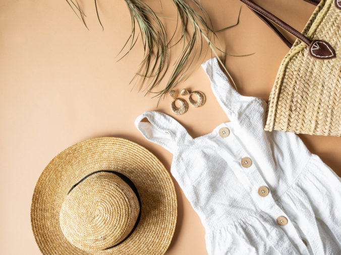 Women&#039;s summer straw hat, wicker bag, white sundress, sunglasses and jewelry on a beige background. Copy space. Top view