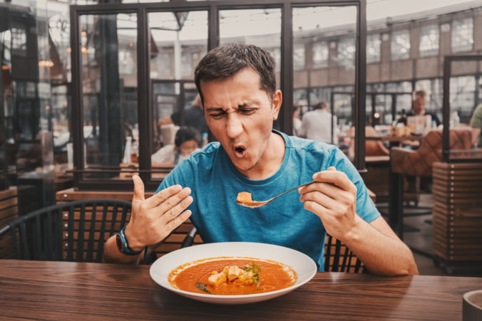 A man tries a spicy and hot red soup in a restaurant and reacts funny emotionally. Seasonings in the national cuisine and an unhealthy diet with overabundance of pepper