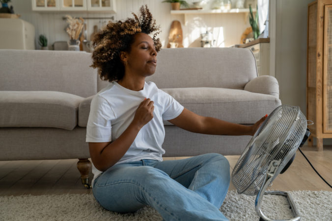 Young African American woman cooling down by ventilator at home, feeling unwell during heatwave