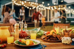 Backyard Dinner Table with Tasty Grilled Barbecue Meat, Fresh Vegetables and Salads. Happy Joyful People Dancing to Music, Celebrating and Having Fun in the Background on House Porch.