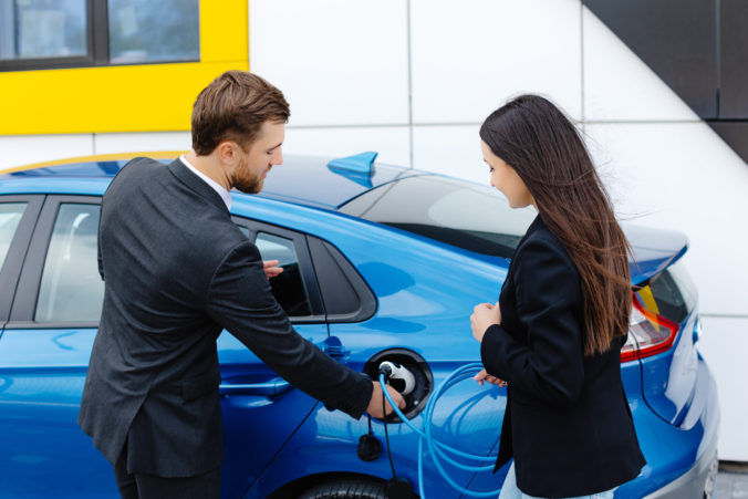 Sales manager showing how to charge electric car to a young client woman, plugging wire into the car socket at the car dealership outdoor