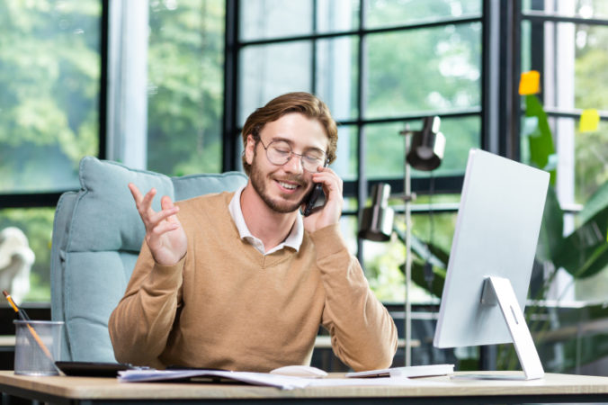 Happy man in the office talking on the phone, sitting at the table in front of the computer, having a successful business conversation, gesturing with his hands