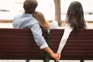 Boyfriend Holding Hands With Girlfriend's Friend Sitting On Bench Outdoor