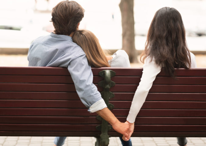 Boyfriend Holding Hands With Girlfriend's Friend Sitting On Bench Outdoor