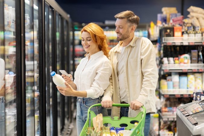 Couple shopping in supermarket together, woman with smartphone scanning bottle of milk, buying groceries, standing with cart in supermarket