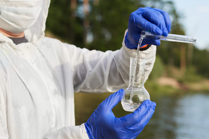 Water Purity Test. Hand in protective gloves holding a chemical flask or test tube with water. Lake in the background.