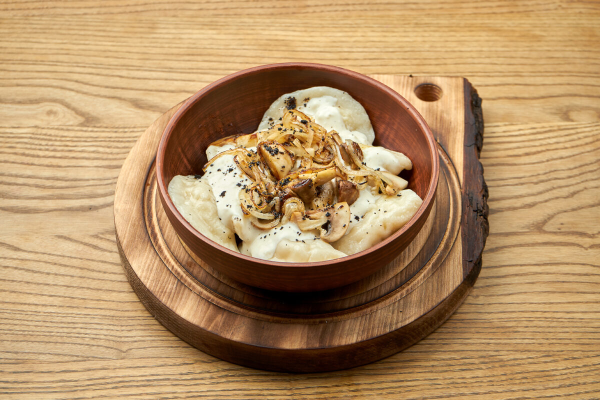 Traditional Ukrainian dumplings filled with mashed potato topping with fried mushroom and sour cream, served in a red bowl on a wooden background. Close up view on Pierogi or varenyky