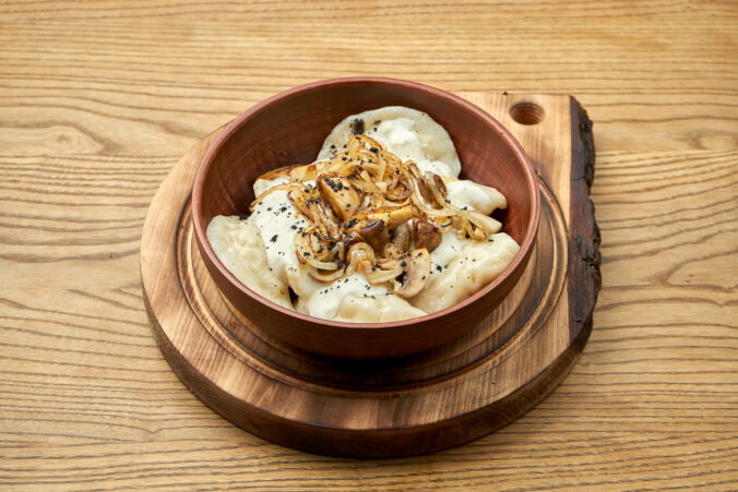 Traditional Ukrainian dumplings filled with mashed potato topping with fried mushroom and sour cream, served in a red bowl on a wooden background. Close up view on Pierogi or varenyky