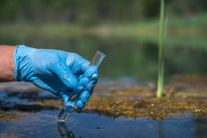 The hand of a working man in a glove holds a test tube with a water sample in close up against the background of a natural landscape.