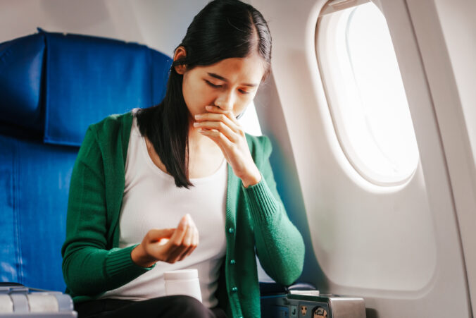 A young Asian woman is sitting in her seat as an airplane passenger, experiencing airsickness with symptoms like nausea, vomiting, dizziness, and general discomfort.